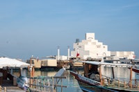 A traditional dhow boat docked at the Museum of Islamic Art in Doha, Qatar, with its distinctive geometric architecture in the background.