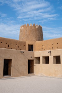 A traditional Qatari desert fort with a prominent round tower and crenelated walls set against a bright blue sky with scattered clouds. The Al Zubara Fort is of significant Qatar cultural heritage.