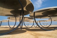 A modern art installation with large, circular structures supported by sleek, curved metal frames on a desert landscape. The Shadows travelling on the sea of the day (2022) created by Olafur Eliasson sits in the Qatari desert.